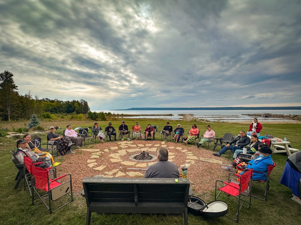 Around 20 students from the Certificate in Journalism in Indigenous Communities Program from Carleton University sitting in lawn chairs around a circular fire pit in front of a large lake, Georgian Bay, with clouds in the sky, green grass surrounding the circle, and trees to their left.